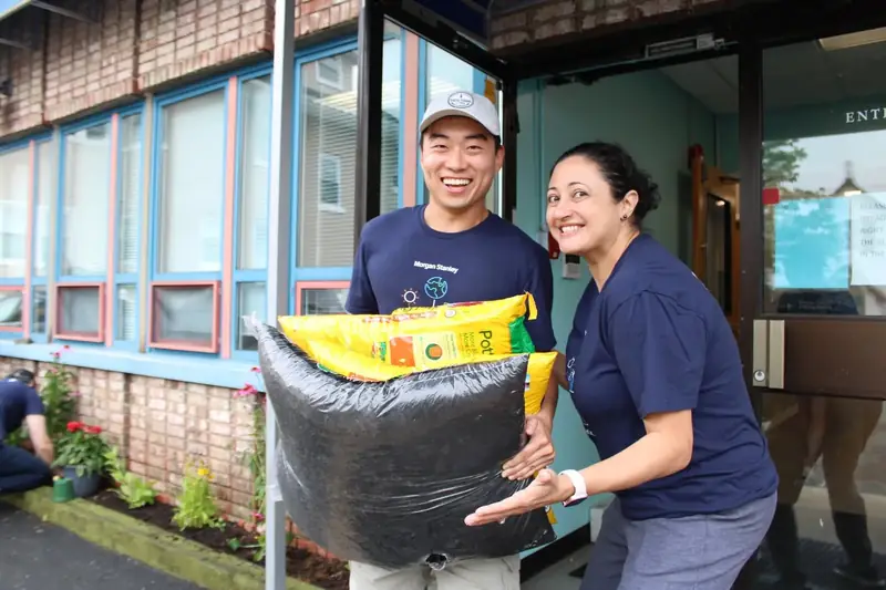 Two smiling volunteers carry bags of soil for the Friendship House garden.